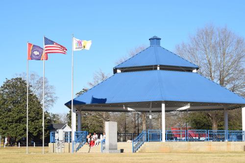 A blue-roofed gazebo with flags in front, surrounded by trees and a grassy area, with people walking nearby.