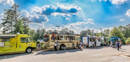A row of colorful food trucks parked under a bright blue sky with fluffy clouds, surrounded by people enjoying the scene.