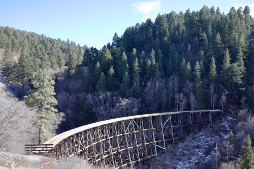 A wooden trestle bridge curves over a valley, surrounded by dense evergreen trees and a clear blue sky.