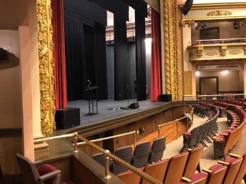A theater stage with red curtains, a microphone stand, and empty seats in a beautifully decorated auditorium.
