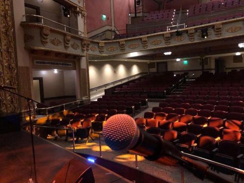A microphone in the foreground with a view of an empty theater, featuring red seats and ornate decor.