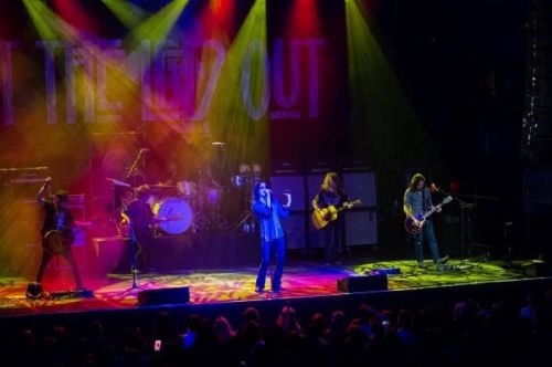 A rock band performs on stage with colorful lights and a lively audience in front.