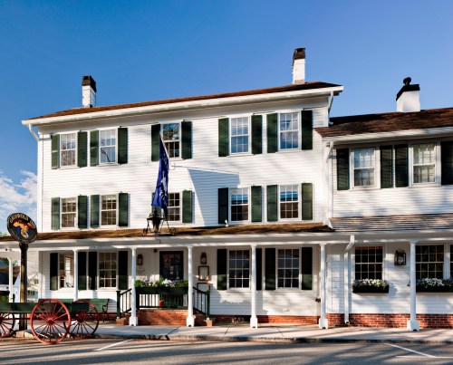 Historic white building with green shutters, a porch, and a sign, set against a clear blue sky.