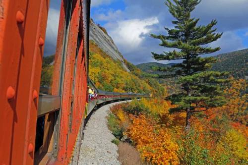 A train curves along a colorful autumn landscape with vibrant trees and a rocky hillside under a cloudy sky.