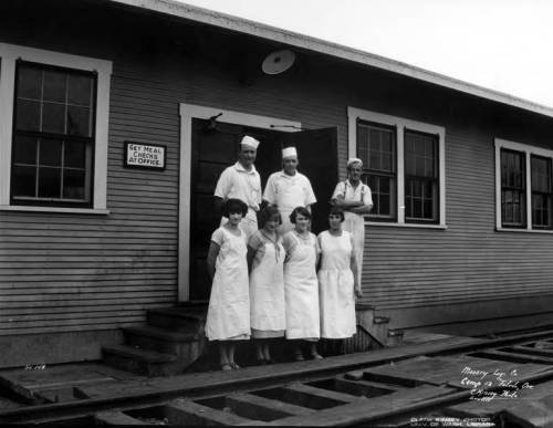 A group of six people in white uniforms stands outside a building, with two men in chef hats and four women in aprons.