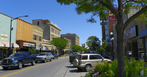 A sunny street scene with shops, parked cars, and trees lining the sidewalk in a small town.