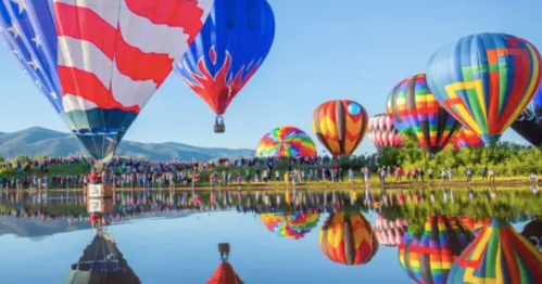 Colorful hot air balloons fill the sky above a crowd by a reflective lake, with mountains in the background.