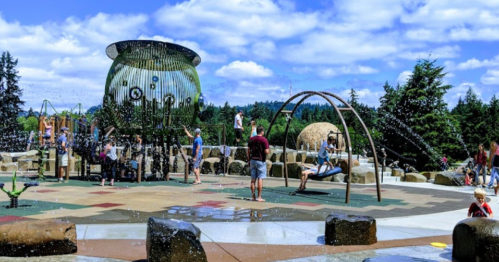 A vibrant playground with water features, swings, and children playing under a blue sky with fluffy clouds.