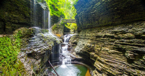 A serene waterfall cascades into a rocky gorge, surrounded by lush greenery and a stone bridge above.