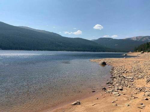 A serene lake surrounded by mountains, with a sandy shore and rocky areas under a clear blue sky.