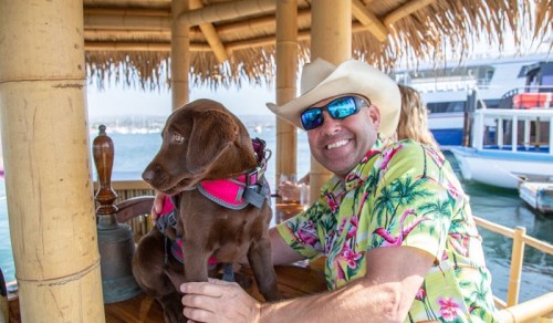 A man in a floral shirt and cowboy hat smiles beside a brown dog wearing a harness on a boat.