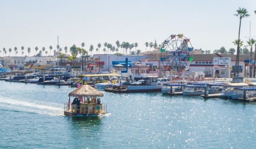 A colorful boat with a thatched roof glides through a sunny harbor, with a ferris wheel and palm trees in the background.