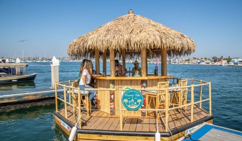 A tiki bar on a floating platform in a marina, with people enjoying drinks under a thatched roof.