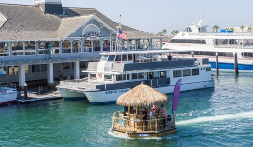 A tiki-style boat floats near a larger ferry at a sunny harbor, with buildings and palm trees in the background.