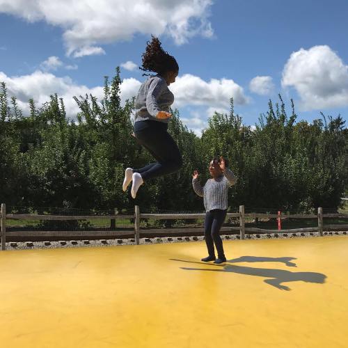 Two children play on a yellow surface, one jumping in the air while the other watches, with trees and blue sky in the background.
