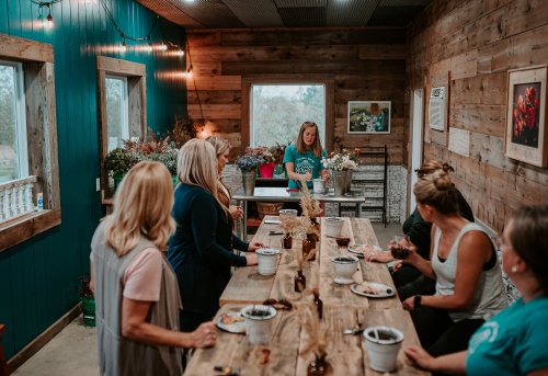 A group of women gather around a rustic wooden table, enjoying food and drinks in a cozy, decorated room.