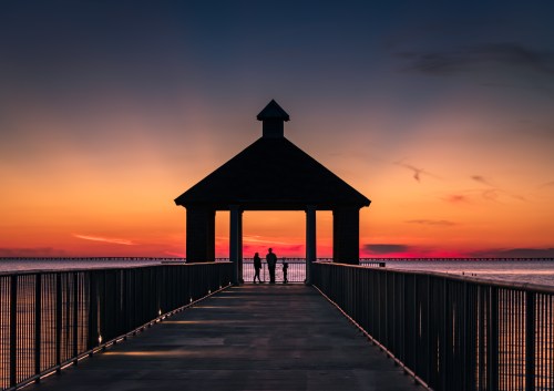 Silhouetted couple standing under a gazebo on a pier at sunset, with vibrant colors in the sky.