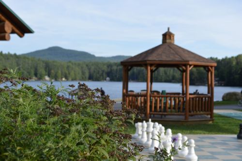 A wooden gazebo by a lake, surrounded by greenery and a large outdoor chess set, with mountains in the background.