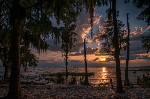 Sunset over a tranquil lake, framed by silhouetted trees and clouds, casting warm reflections on the water.