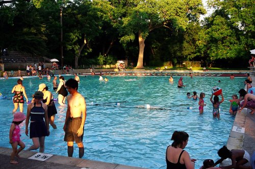 A busy outdoor swimming pool filled with people enjoying the water on a sunny day, surrounded by trees.