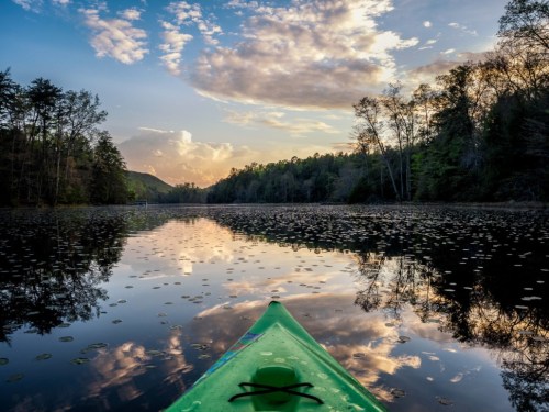 A green kayak on calm water reflects clouds and trees at sunset, creating a serene natural landscape.