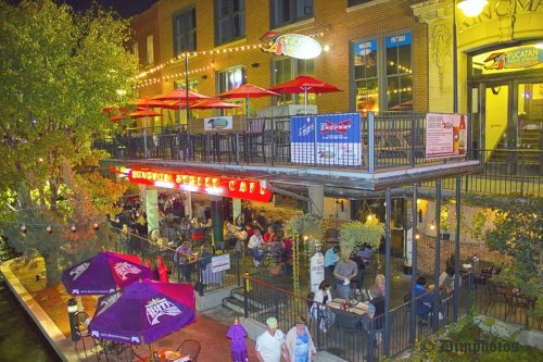 A lively outdoor café at night, with diners enjoying meals under string lights and colorful umbrellas along a canal.