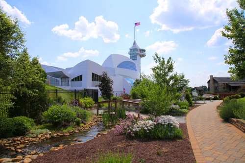 A modern building with a flag, surrounded by greenery, a stream, and a pathway on a sunny day.