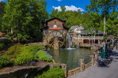 A scenic view of a watermill beside a pond, surrounded by greenery and buildings under a clear blue sky.