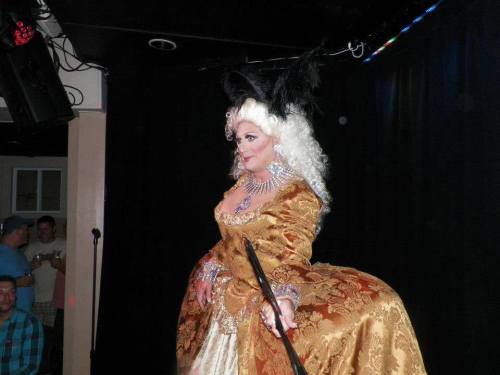 A performer in a lavish gold gown with feathers, striking a pose on stage, showcasing a dramatic look.