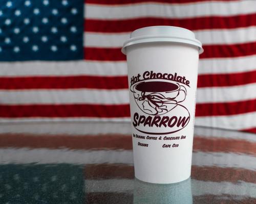 A white coffee cup with "Hot Chocolate Sparrow" logo in front of an American flag backdrop.
