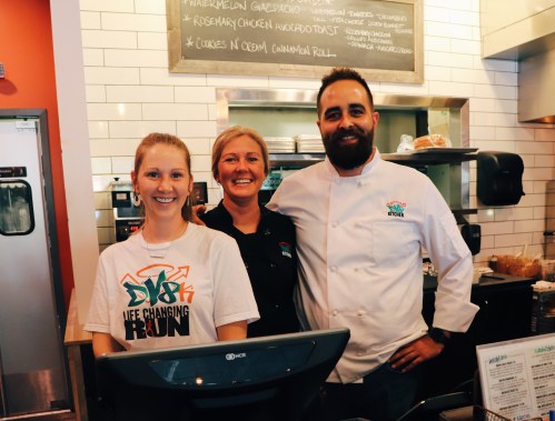 Three smiling staff members stand behind a counter in a cafe, with a menu board and food items in the background.