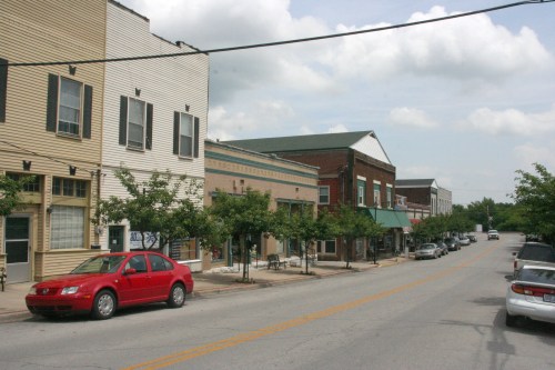 A quiet street lined with shops and trees, featuring a red car parked along the curb under a cloudy sky.