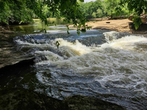 A serene river scene with flowing water, surrounded by lush greenery and rocky banks under a clear blue sky.