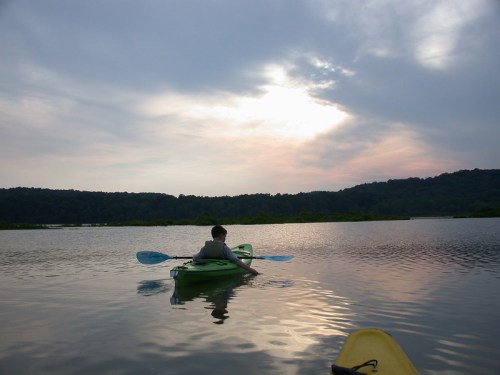A person kayaking on a calm lake at sunset, with hills in the background and a cloudy sky.