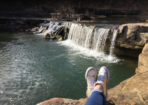 A person’s feet in sneakers resting on a rock by a serene waterfall and calm water.