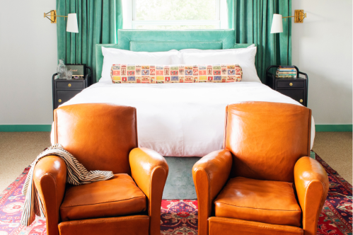 A cozy bedroom featuring a large bed with a decorative pillow and two brown leather chairs in front on a patterned rug.