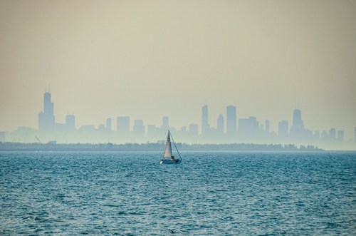 A sailboat glides across a calm lake with a hazy city skyline in the background.