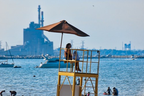 A lifeguard sits under an umbrella on a tall chair by the beach, with boats and people enjoying the water in the background.