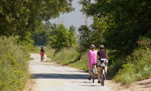 Two cyclists ride on a gravel path surrounded by greenery, while another cyclist approaches from behind.