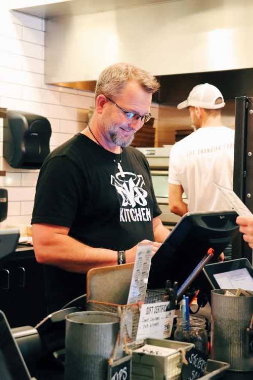 A smiling man at a restaurant counter checks a device while another staff member works in the background.