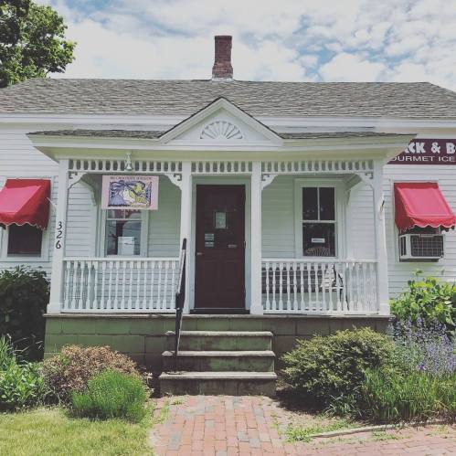 A charming white house with a porch, red awnings, and a sign for gourmet ice cream. Lush greenery surrounds the entrance.