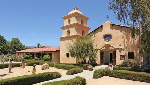A charming adobe-style building with a bell tower, surrounded by greenery and sculptures in a sunny outdoor setting.