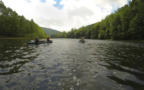 Two people in kayaks on a calm river surrounded by lush green trees and mountains under a partly cloudy sky.