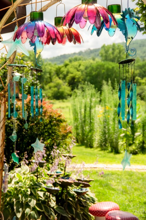 Colorful glass wind chimes and decorations hang from a tree, set against a lush green garden backdrop.