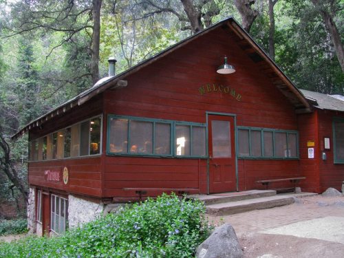 A rustic red cabin surrounded by trees, featuring large windows and a welcoming sign above the entrance.