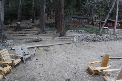 A rustic outdoor area with wooden chairs, stone walls, and a cabin surrounded by trees in a forest setting.