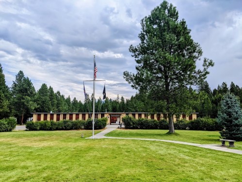A grassy area with trees and a building in the background, featuring flags and a cloudy sky.