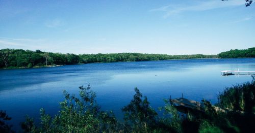 A serene lake surrounded by lush greenery under a clear blue sky, with a small dock visible at the water's edge.