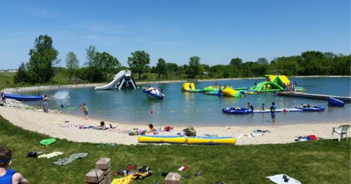 A sunny beach scene with children playing in a lake, inflatable slides, and water toys, surrounded by green trees.
