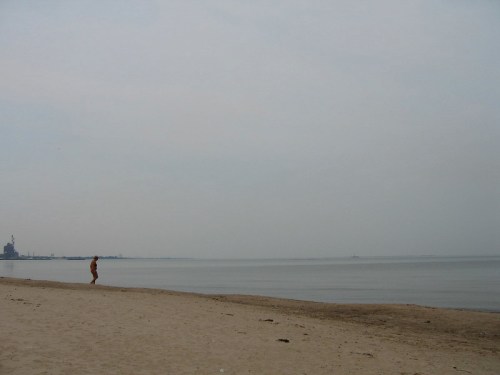A person walks along a sandy beach, with calm water and a hazy sky in the background.